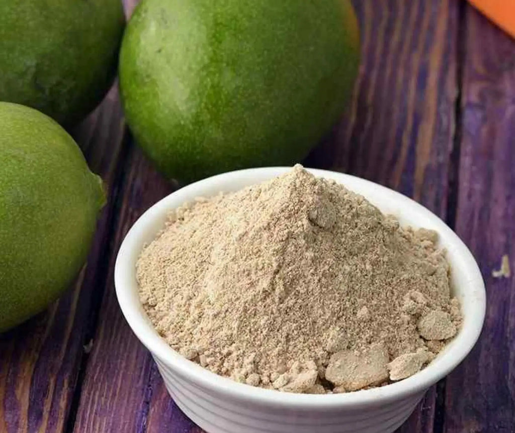 White bowl filled with brown vedcare amchur powder next to green fruits on a wooden surface